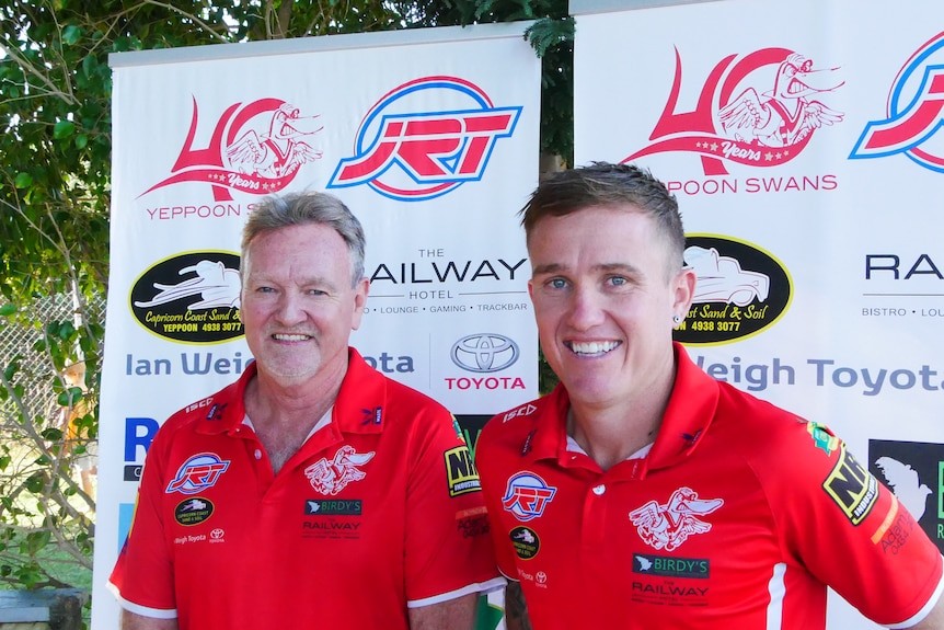 Two men smiling wearing red Yeppoon Swans uniforms in front of trees and a banner