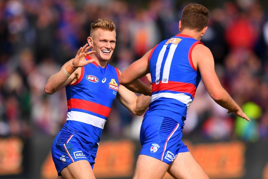 A Western Bulldogs AFL player smiles as he runs towards a teammate to congratulate him on kicking a goal.