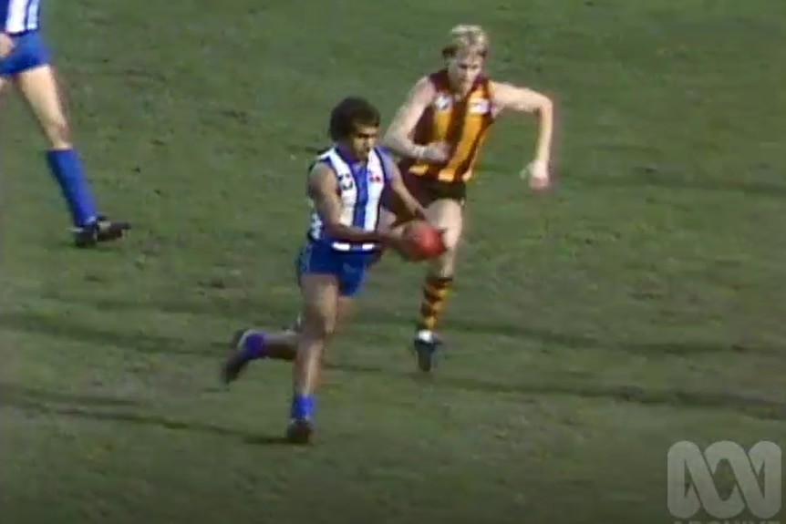 North Melbourne footy player running with ball with Hawthorn player chasing