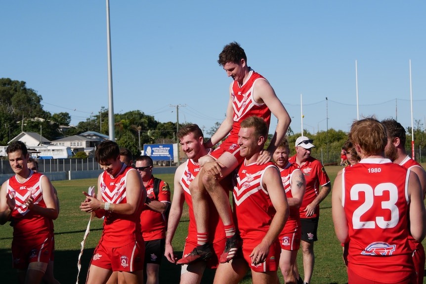 Two smiling AFL players in red and white singlets hoist another player onto their shoulders.