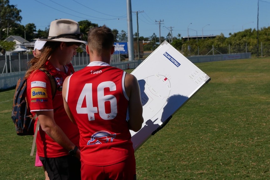 A man with a hat holding a board as another man in an AFL uniform looks on and talks with him