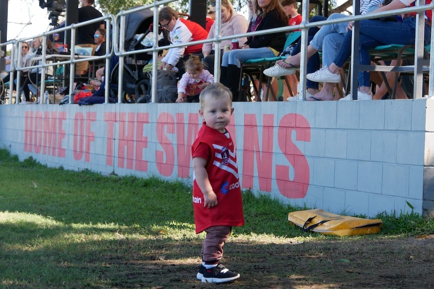 A young boy in a red and white AFL uniform in front of a grand stand with the word Home of the Swans painted on it