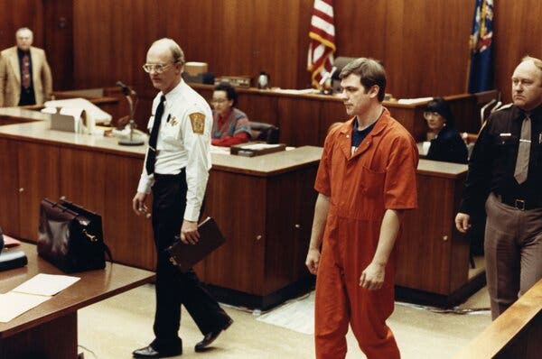 Jeffrey Dahmer, in an orange prison uniform, in a courtroom. His arms are at his sides and there are court officers on either side of him.