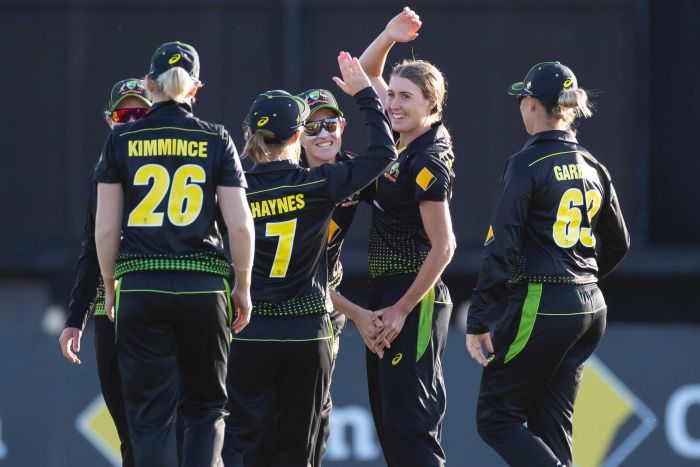 A female cricketer raises her right arm as she celebrates with her teammates, who are surrounding her.