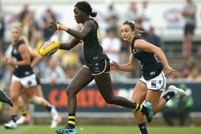 A woman dressed in an AFL uniform and football boots runs with an AFL ball while being chased by another woman.