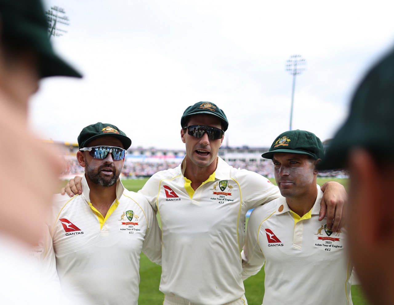 Three Australian cricketers stand huddled beside one another on the pitch.